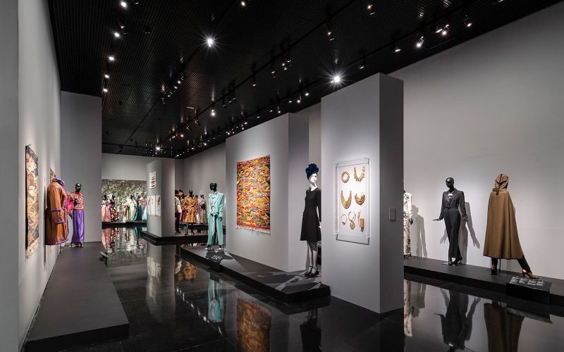 L'image montre l'intérieur minimaliste d'une galerie du musée Yves Saint Laurent à Marrakech, où des mannequins élégants sont mis en valeur par un éclairage tamisé, créant une ambiance de sophistication et d'élégance. Cette exposition met en avant les créations emblématiques du couturier, offrant aux visiteurs une immersion dans l'univers de la haute couture.