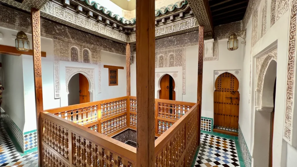 La grande cour centrale de la médersa Ben Youssef est ornée d'un bassin rectangulaire, entouré de colonnes en marbre et de murs décorés de zellige coloré, mettant en valeur la splendeur de l'architecture islamique à Marrakech. Ce lieu historique, véritable joyau du patrimoine mondial, invite les visiteurs à découvrir son ambiance paisible et son importance dans l'enseignement théologique.
